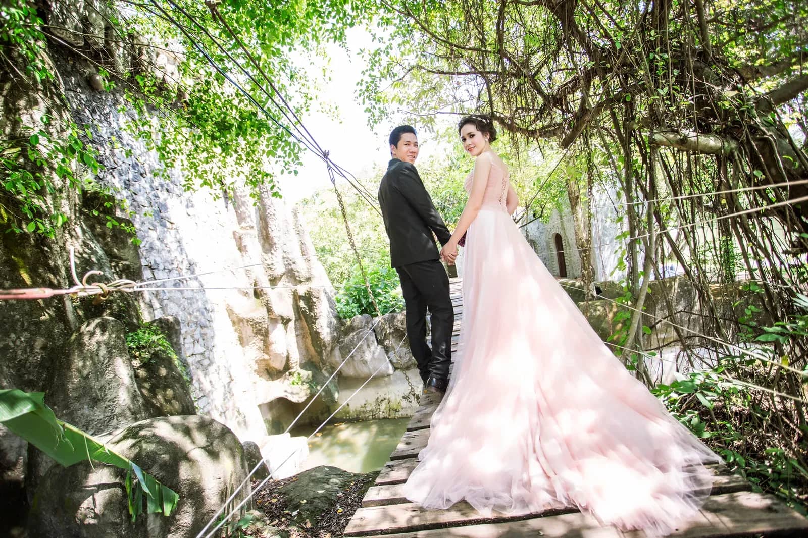 Cô dâu chú rể trên cầu gỗ bên tường đá và dòng nước chảy